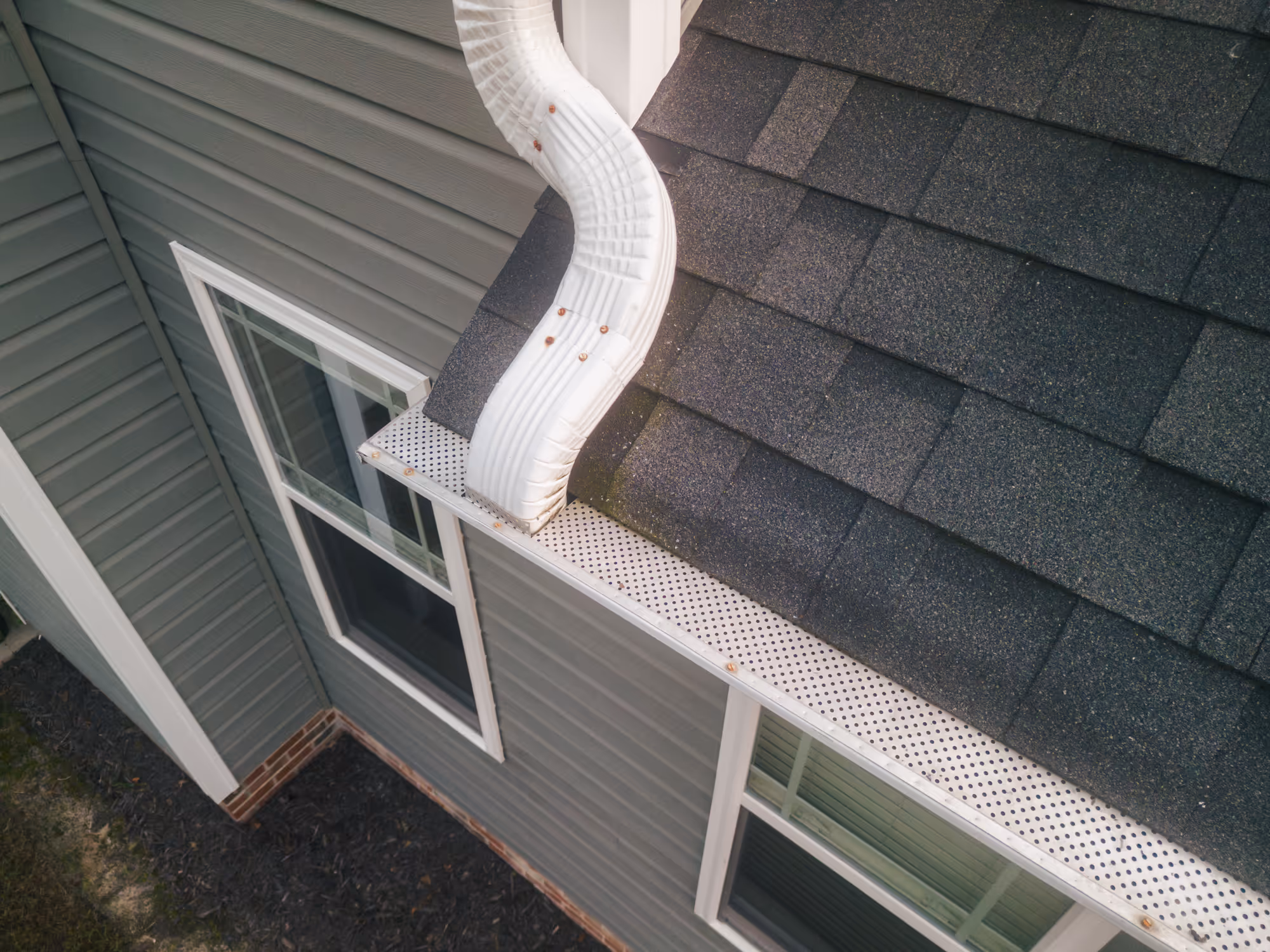 An overhead view of a perforated gutter guard system installed along a residential roofline with a white downspout extending from the gutters above a window.